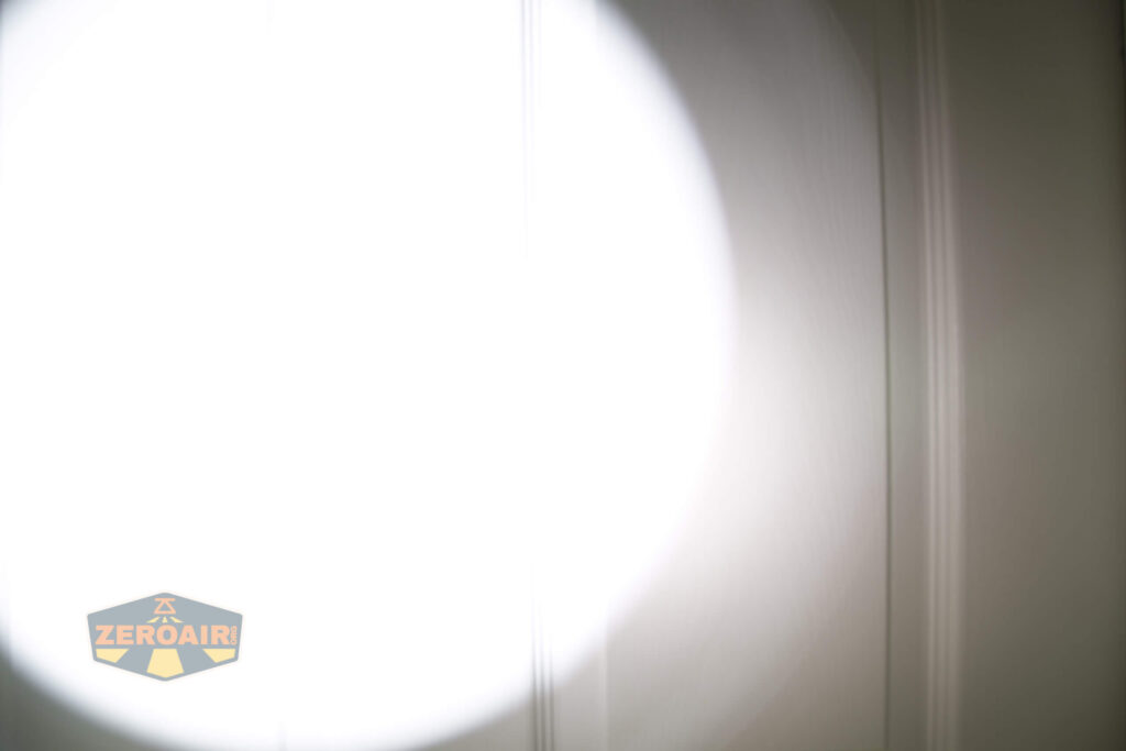 A white door with vertical panels is illuminated by a bright, circular beam of light, likely from a flashlight, causing most of the left side of the image to appear overexposed. The ZeroAir logo is in the bottom left corner.