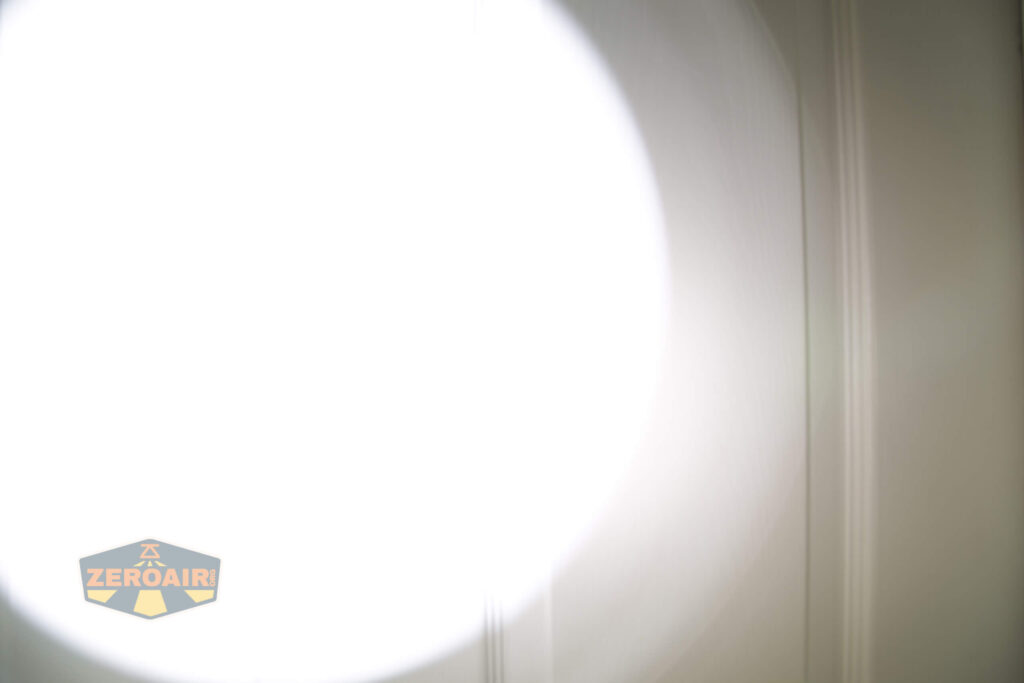 A bright white flashlight beam is cast on a plain wall, creating a circular light pattern. The wall features vertical lines, and a ZEROAIR watermark appears in the bottom left corner.