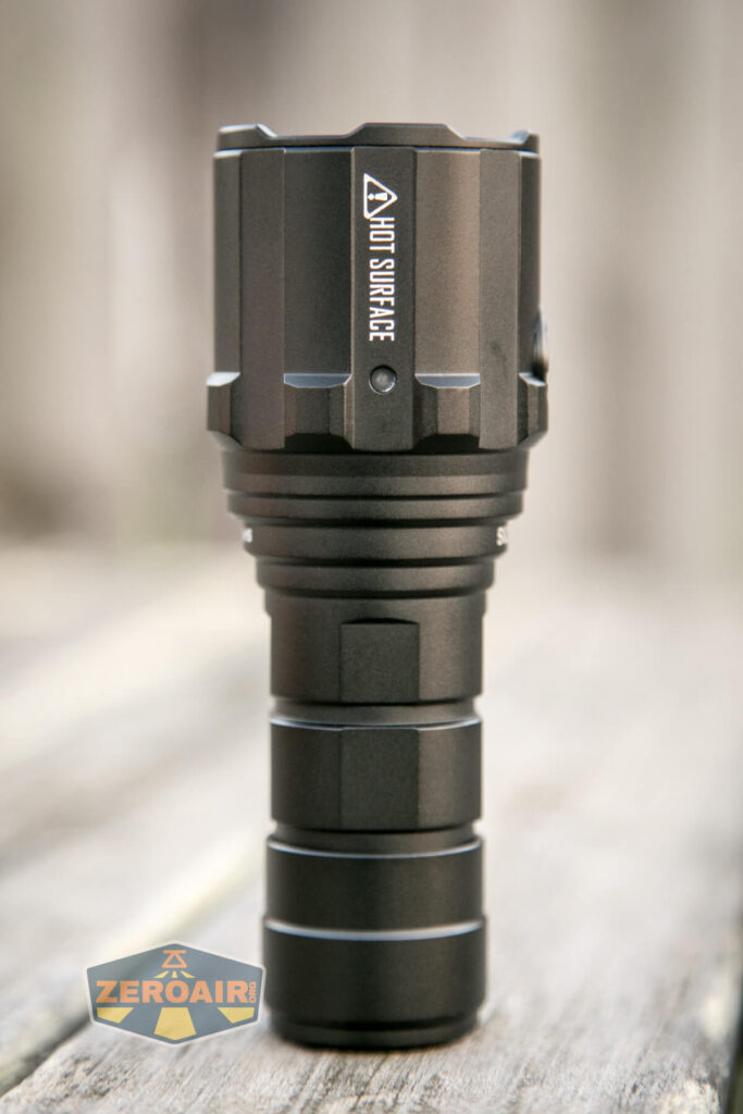 A black, cylindrical flashlight with a warning label HOT SURFACE on the head, standing upright on a wooden surface. The logo ZEROAIR appears in the lower left corner.