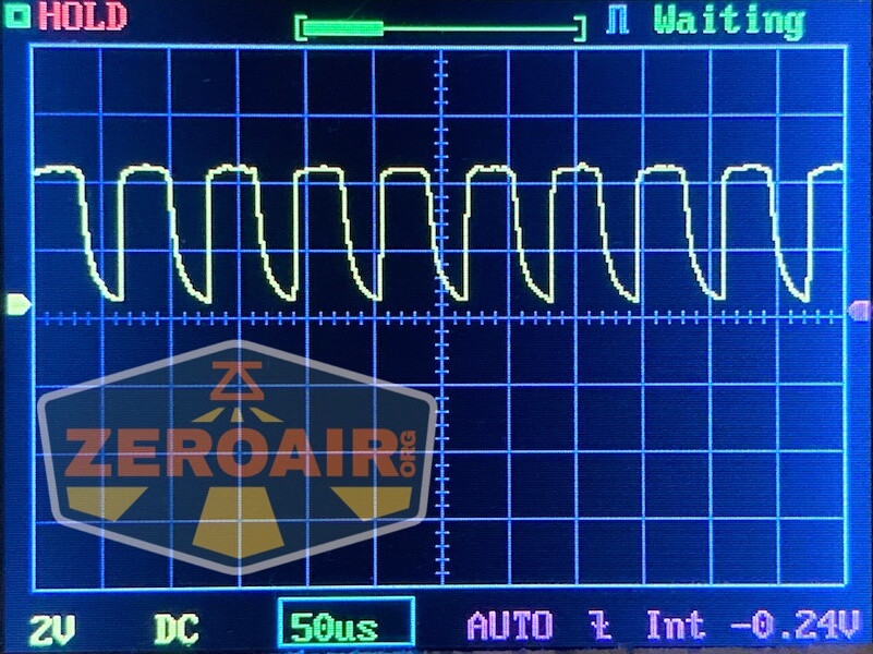 An oscilloscope screen displays a repeating, irregular waveform in yellow. The gridlines, voltage and time settings are visible, alongside a Lumintop GT Nano Brass resting nearby and a large ZeroAir logo overlay in the lower left corner.