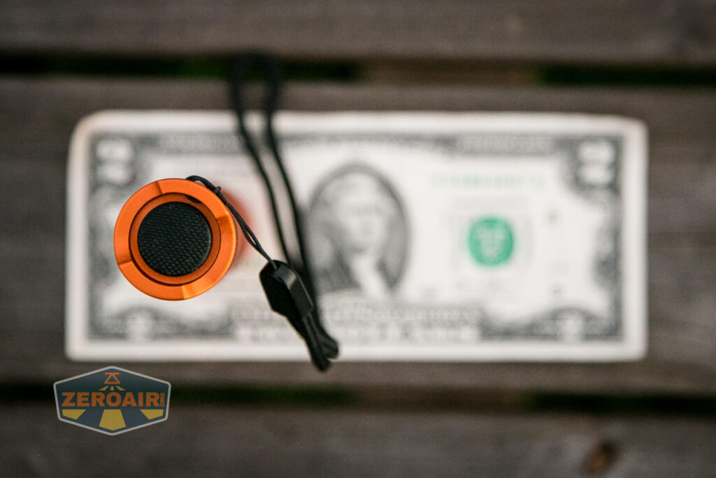 An orange flashlight with a black button and wrist strap is positioned vertically above a US dollar bill on a wooden surface. A logo with the text ZEROAIR is visible in the bottom left corner.