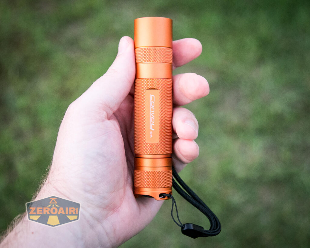A hand holding an orange Convoy S2+ flashlight with a wrist strap, against a blurred green outdoor background. The Zeroair logo appears in the bottom left corner.