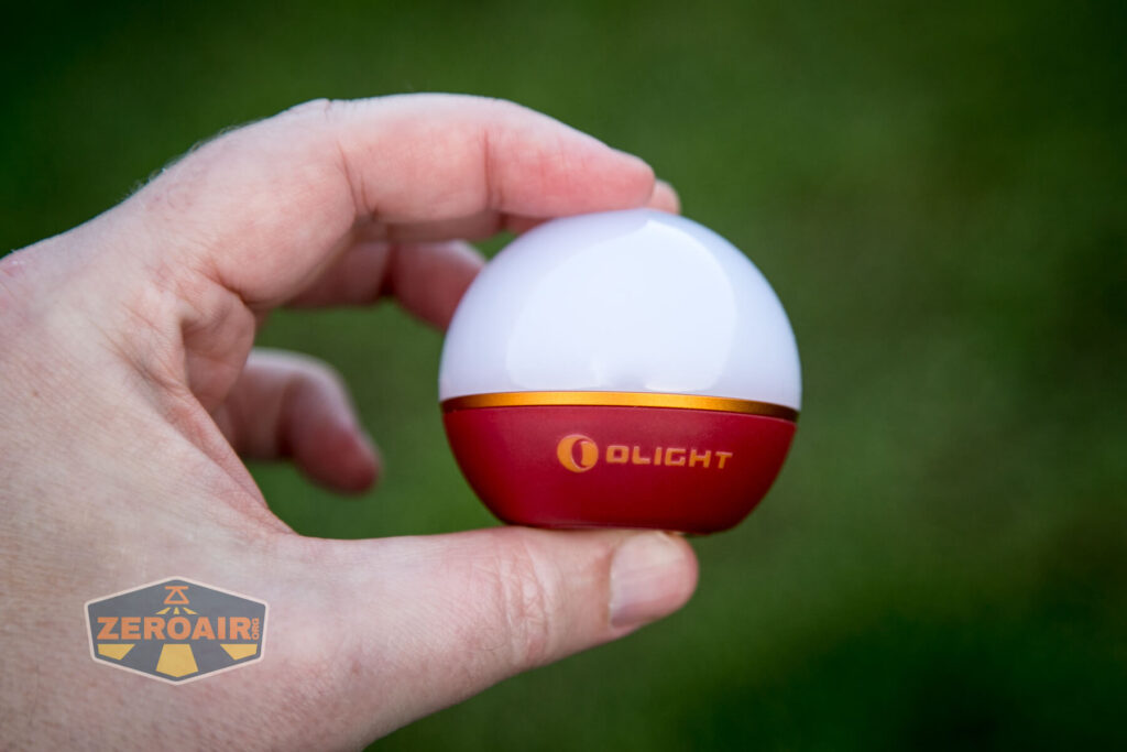 A hand holds a small, spherical object with a white top and red base labeled Olight in gold lettering. The background is blurred green, and a ZeroAir logo appears in the lower left corner.