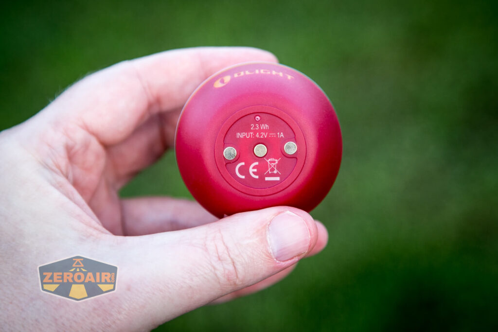 A hand holds a small, round red device labeled “OLIGHT” with technical specifications and certification symbols printed on the bottom against a blurred green background. The ZeroAir logo is in the lower left corner.