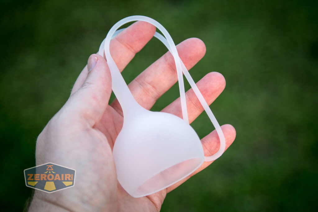 A hand holds a translucent, flexible silicone cap with an attached loop. The background is out of focus green grass. A watermark in the lower left corner features the text ZEROAIR.