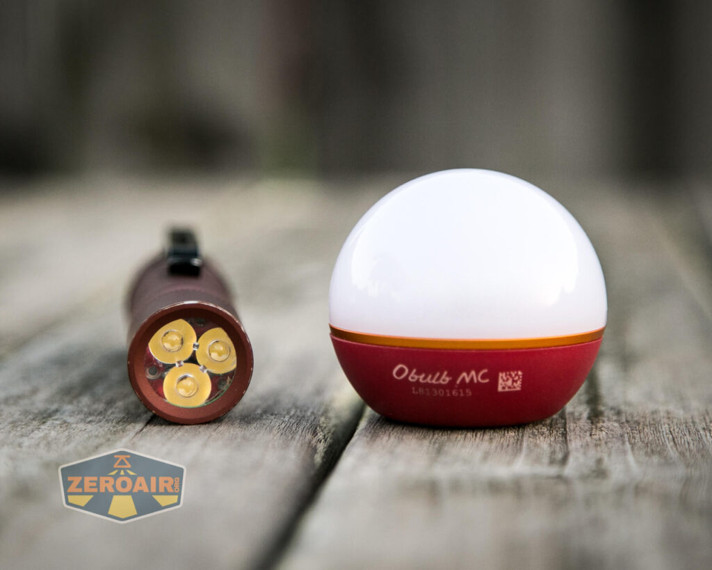 A small flashlight with three LED bulbs lies on a wooden surface next to a round, red and white object marked Olight MC. A ZeroAir logo is visible in the lower left corner.