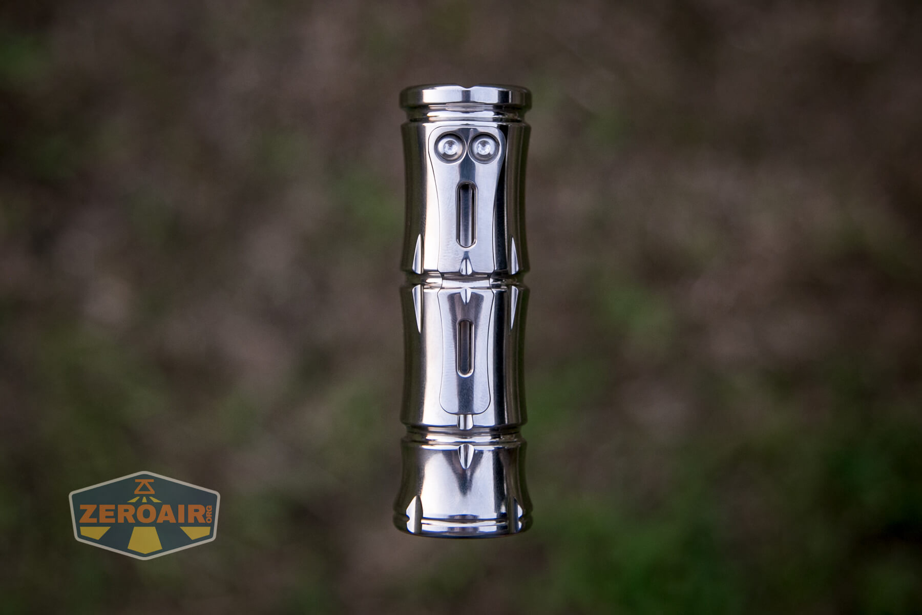 A shiny, metallic cylindrical object with a face-like design stands upright against a blurred brown and green background. The ZeroAir logo is visible in the bottom left corner.