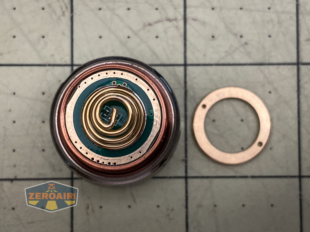 Close-up of a circular electronic component with a coiled gold wire on a green and copper circuit board, the WWLZ Bamboo Small copper ring beside it, all resting on a grid-patterned surface with a ZEROAIR logo.