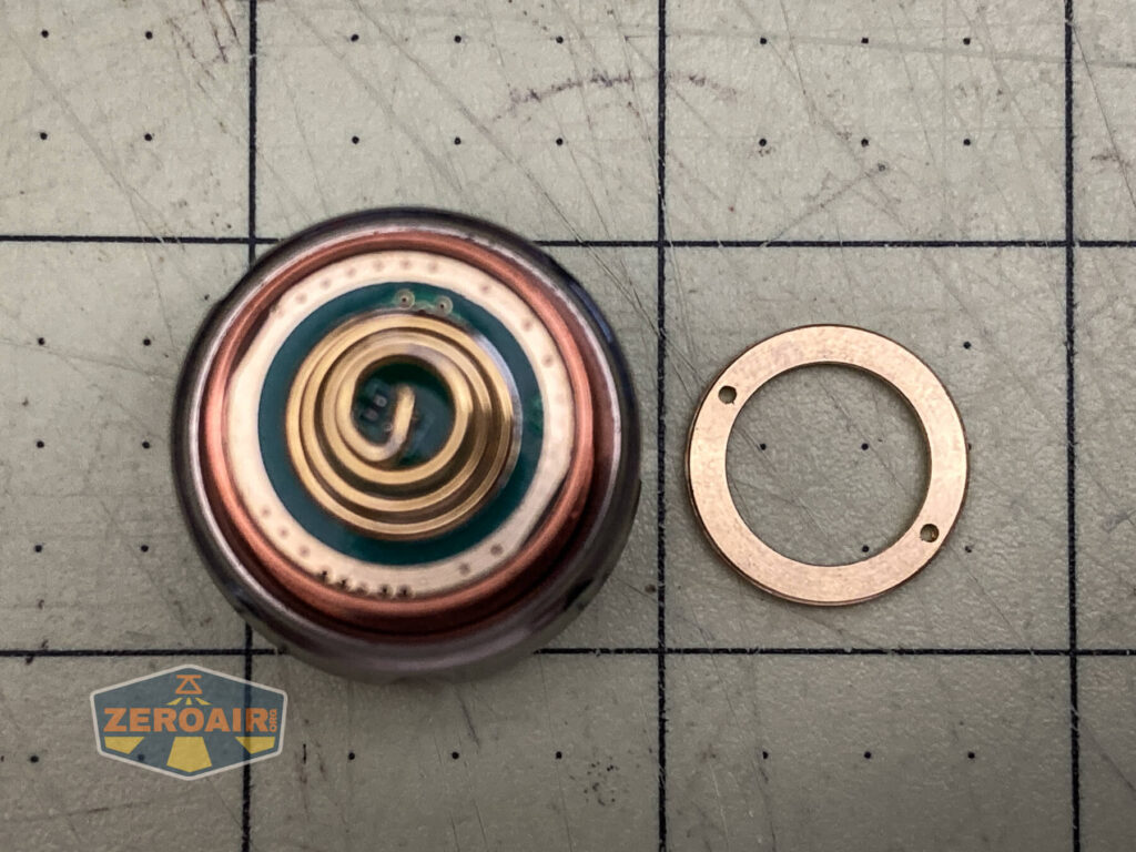 A close-up of a circular electronic component with concentric metal rings and an adjacent thin copper ring, placed on a gridded cutting mat next to a WWLZ Bamboo Small. A ZeroAir logo appears in the corner.