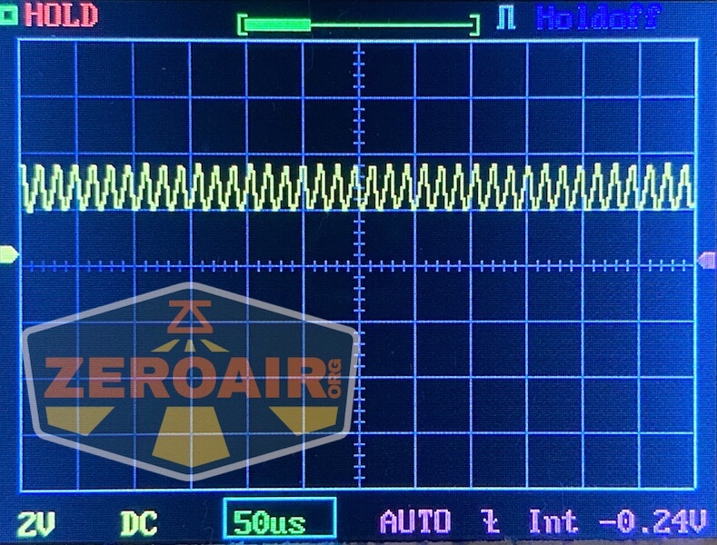 An oscilloscope screen displays a repeating waveform signal in yellow. The settings at the bottom show 2V per division, 50 microseconds per division, and a measured value of -0.24V. The ZEROAIR logo is overlaid.