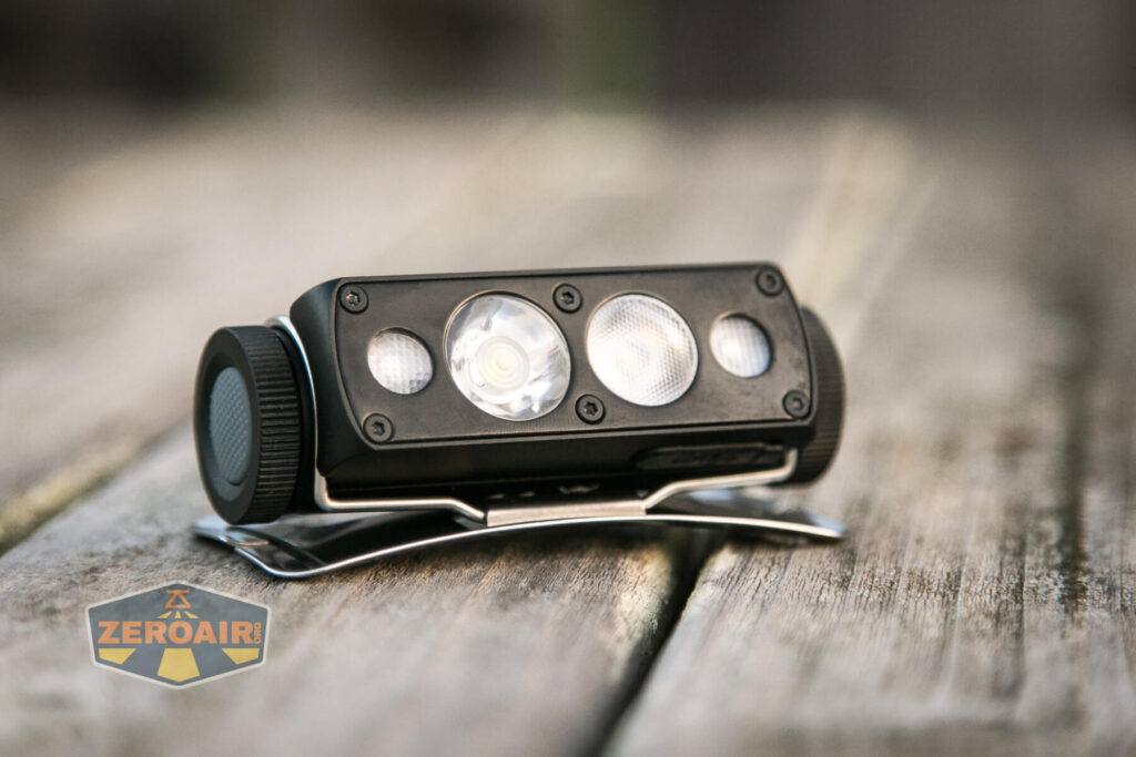 A black, rectangular flashlight with three LED lenses sits on a metal stand on a wooden surface. The logo ZEROAIR is visible in the lower left corner of the image.