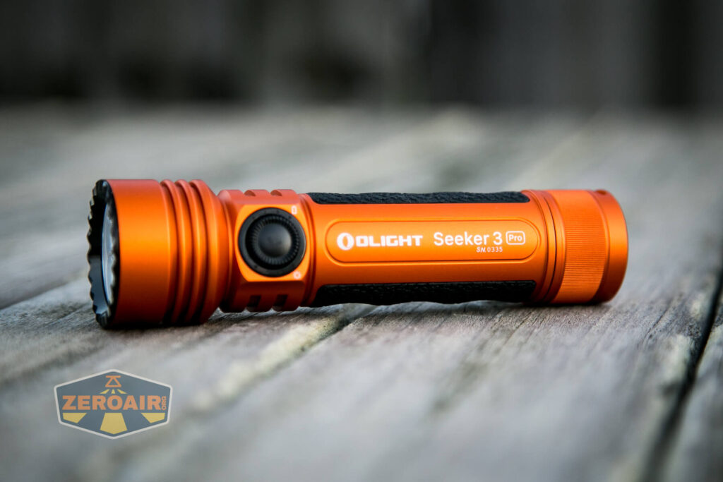 An orange Olight Seeker 3 Pro flashlight rests on a wooden surface. The flashlight features a black rubber grip, a round black button, and branding on the side. The logo ZeroAir is visible in the lower left corner.