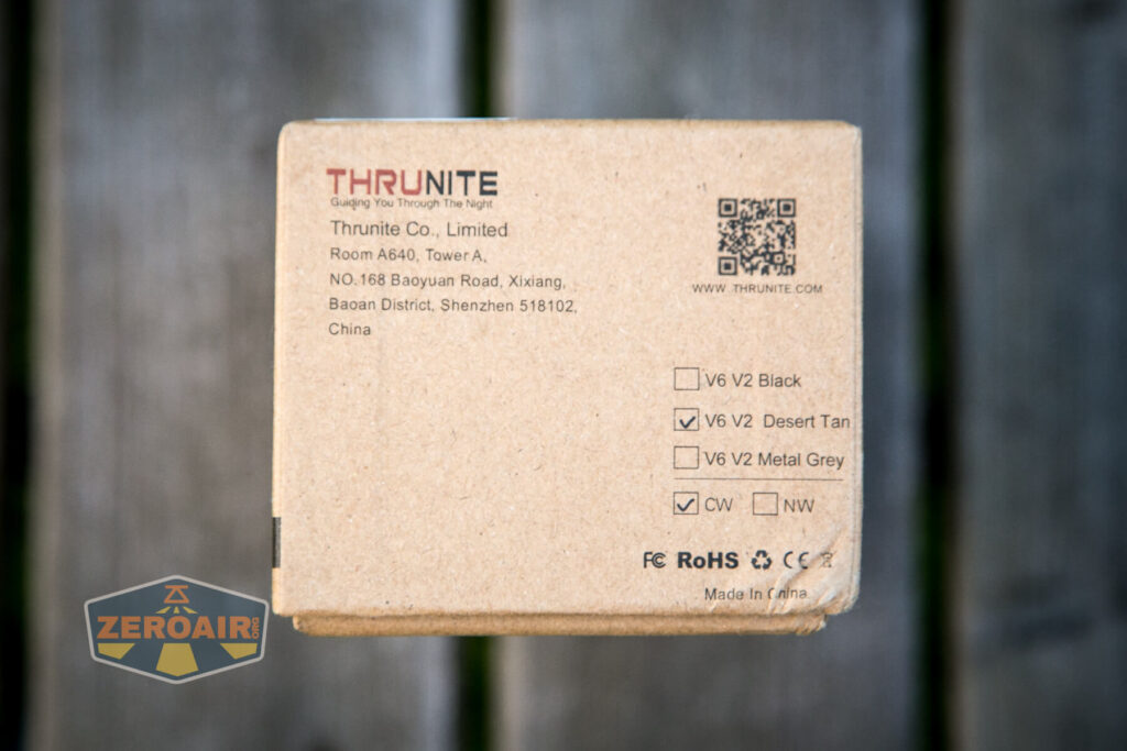 A cardboard box from ThruNite Co., Limited displays the company address, product options for V6 V2 in Black, Desert Tan, and Metal Grey, with Metal Grey checked. A QR code and certification marks are also visible.