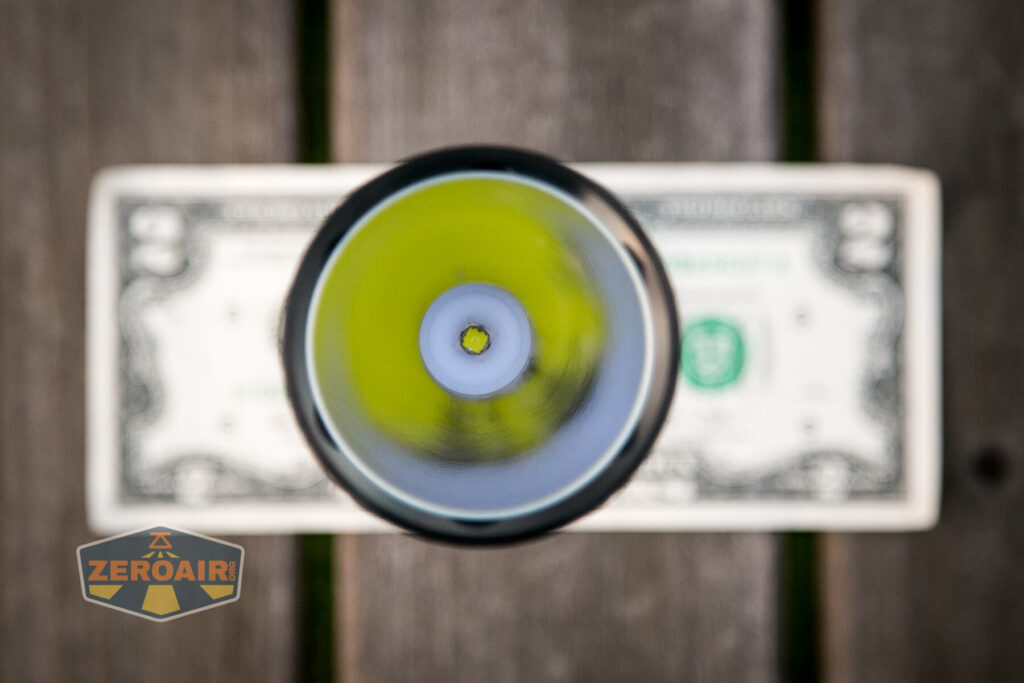 A close-up, top-down view of a flashlight lens centered over a U.S. two-dollar bill on a wooden surface; the flashlight emits a yellowish glow. A ZeroAir logo is visible in the bottom left corner.