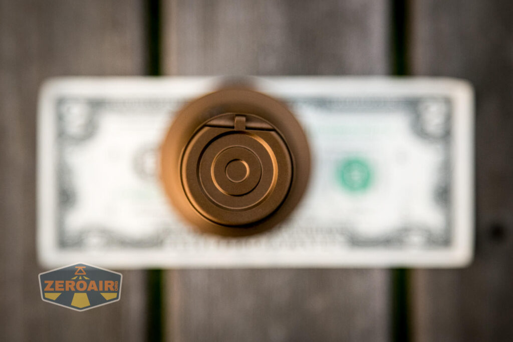 A bronze-colored cylindrical object viewed from above is centered over a blurred US two-dollar bill, with a ZeroAir logo in the bottom left corner. The background appears to be wooden planks.