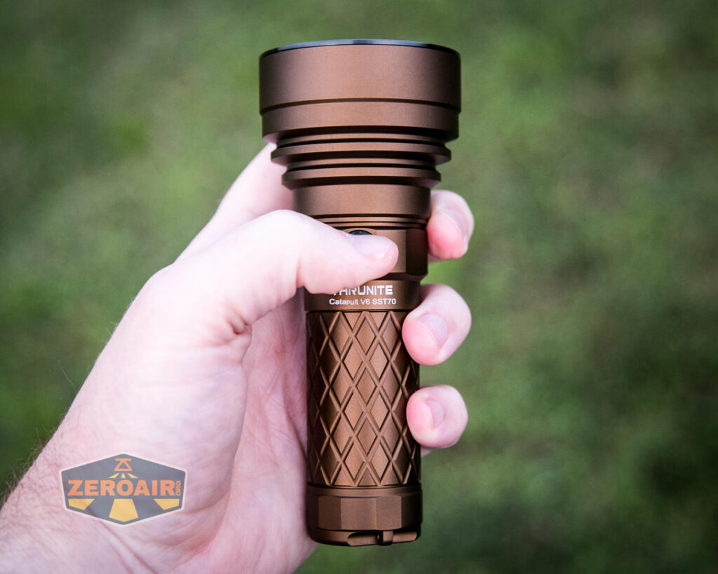 A hand holding a bronze-colored flashlight with a textured grip, viewed against a blurred green background. The bottom left corner displays a logo that says ZeroAir.