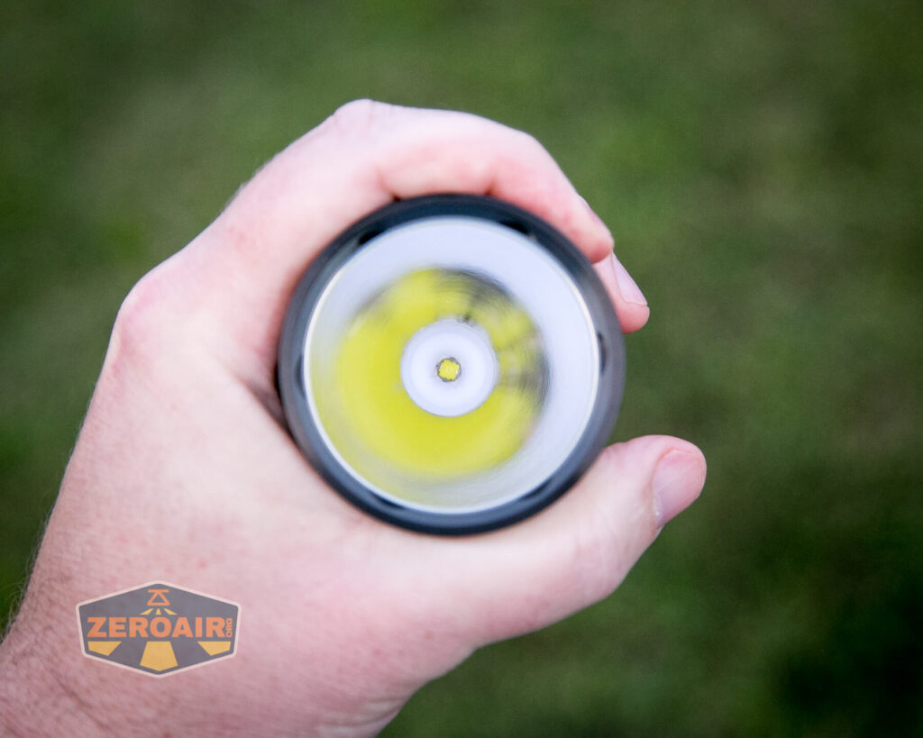 A hand holding a flashlight pointed toward the camera, showing the flashlight’s LED bulb and reflector. The background is blurred green grass. A ZeroAir logo appears in the lower left corner.