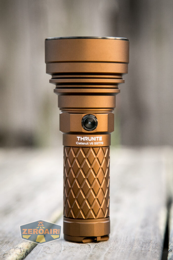 A copper-colored ThruNite Catapult V6 SST70 flashlight stands upright on a wooden surface, featuring a textured grip and a single black button on the side. The ZeroAir logo is partially visible in the bottom left corner.