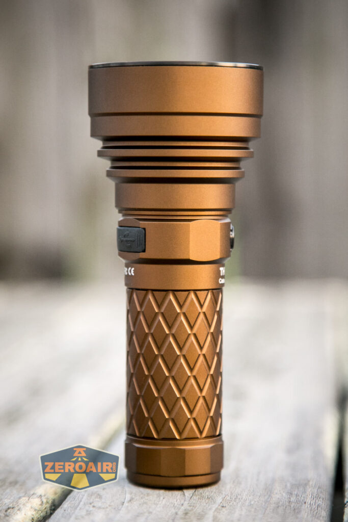 A bronze-colored flashlight with a textured, diamond-patterned grip stands upright on a wooden surface. The background is blurred, and a ZeroAir logo appears in the lower-left corner.