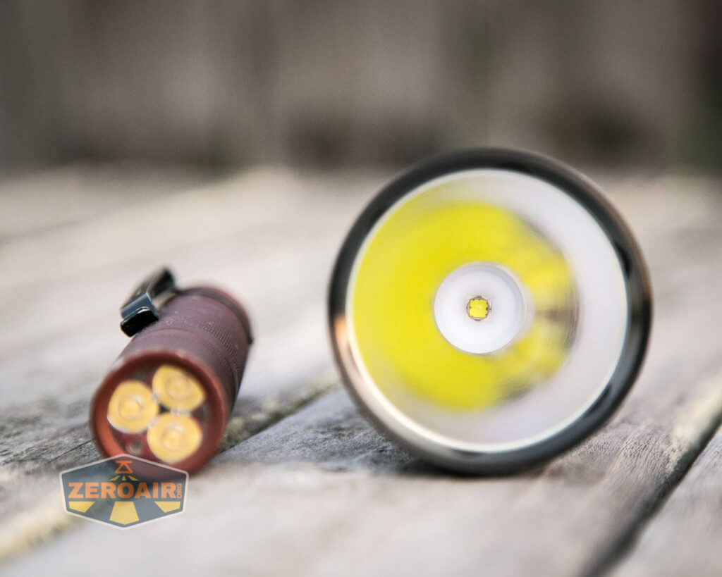 A close-up of two flashlights on a wooden surface; the left flashlight shows its three LED bulbs, while the right flashlight displays its large, single LED and reflector facing the camera.
