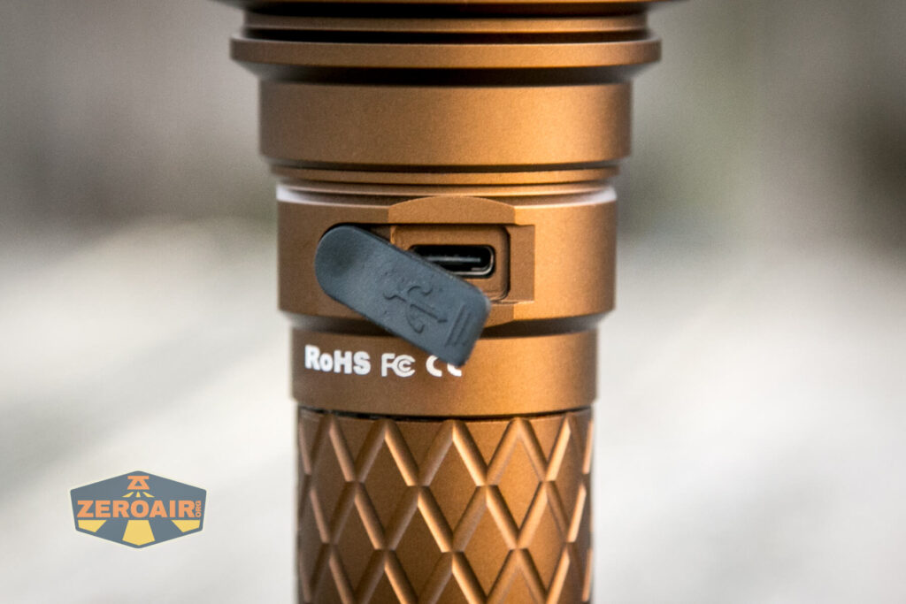 Close-up of a metallic flashlight with a diamond-patterned grip and a rubber cover open to reveal a USB-C charging port. RoHS and FCC certification marks are visible. A Zeroair logo appears in the lower left corner.