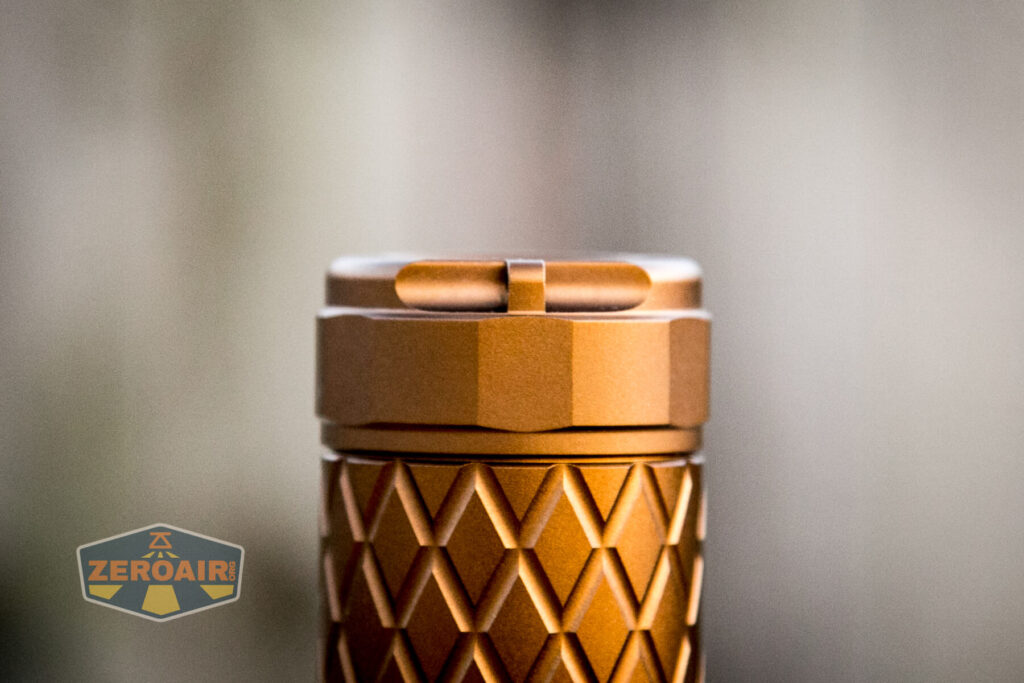 Close-up of the top of a cylindrical, copper-colored object with a textured diamond pattern and flat top. The background is blurred. The lower left corner contains a logo with the word ZEROAIR.