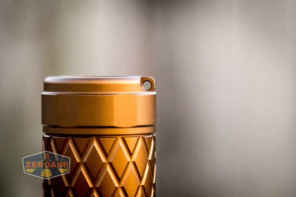 Close-up of the top section of a bronze-colored, textured cylindrical object with a small hole on the lid, positioned against a blurred background. The ZEROAIR logo appears in the lower left corner.