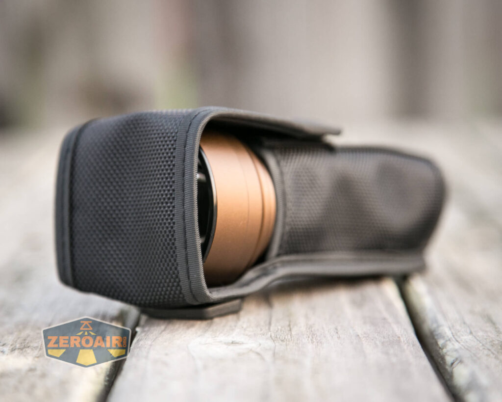 A bronze flashlight partially enclosed in a black textured fabric case rests on a wooden surface. The case flap is open, revealing the flashlight’s head. A ZeroAir logo is visible in the bottom left corner.