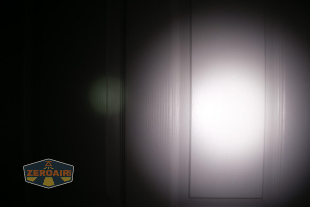 A white flashlight beam shines on a white door in a dark room, creating a bright circular spot in the center and a faint green spot to the left. The ZeroAir logo appears in the bottom left corner.