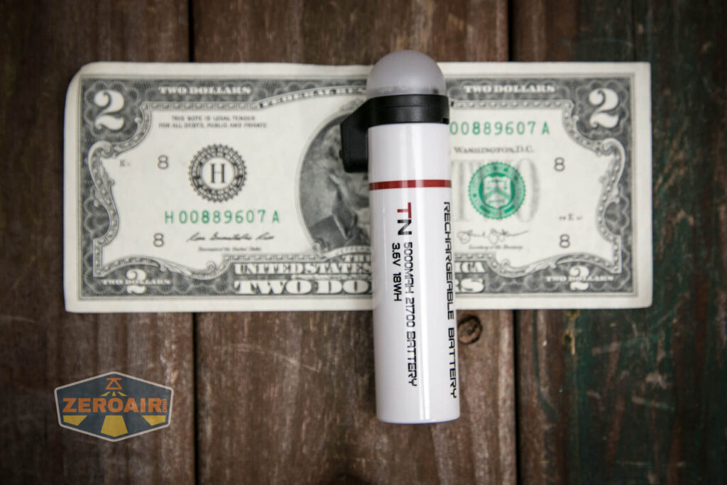 A cylindrical rechargeable battery labeled T:N 30SF 3.7V 3000mAh stands upright on a two-dollar bill, which rests on a wooden surface. A ZeroAir logo is in the bottom left corner.