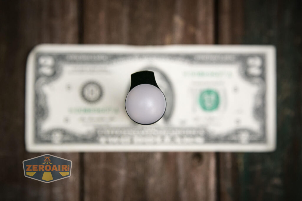 A cylindrical object with a white rounded top is standing upright on a U.S. two-dollar bill, placed on a wooden surface. The ZeroAir logo appears in the lower left corner.