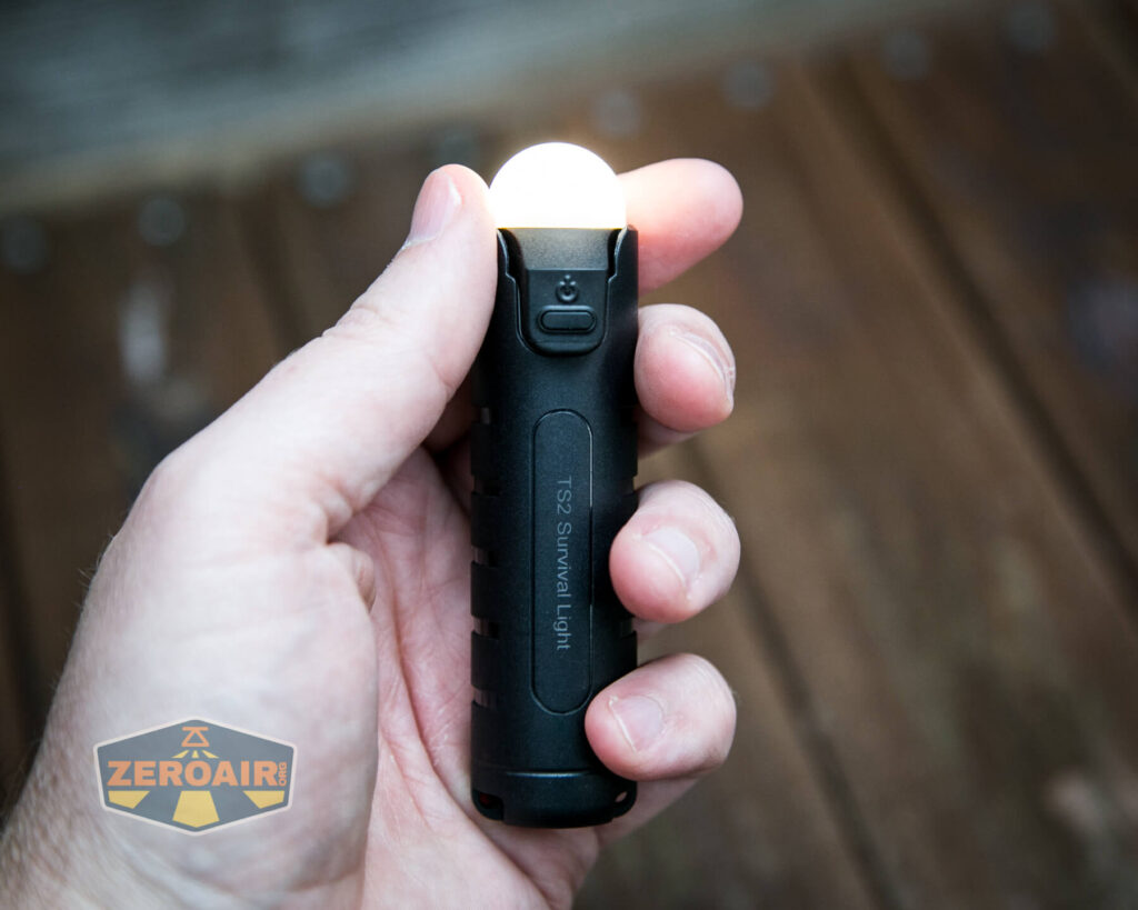 A hand holding a small black flashlight with a glowing white light. The flashlight has ridges and a switch near the light. TS2 Survival Light is labeled on the side. A wooden surface and ZeroAir logo are visible in the background.