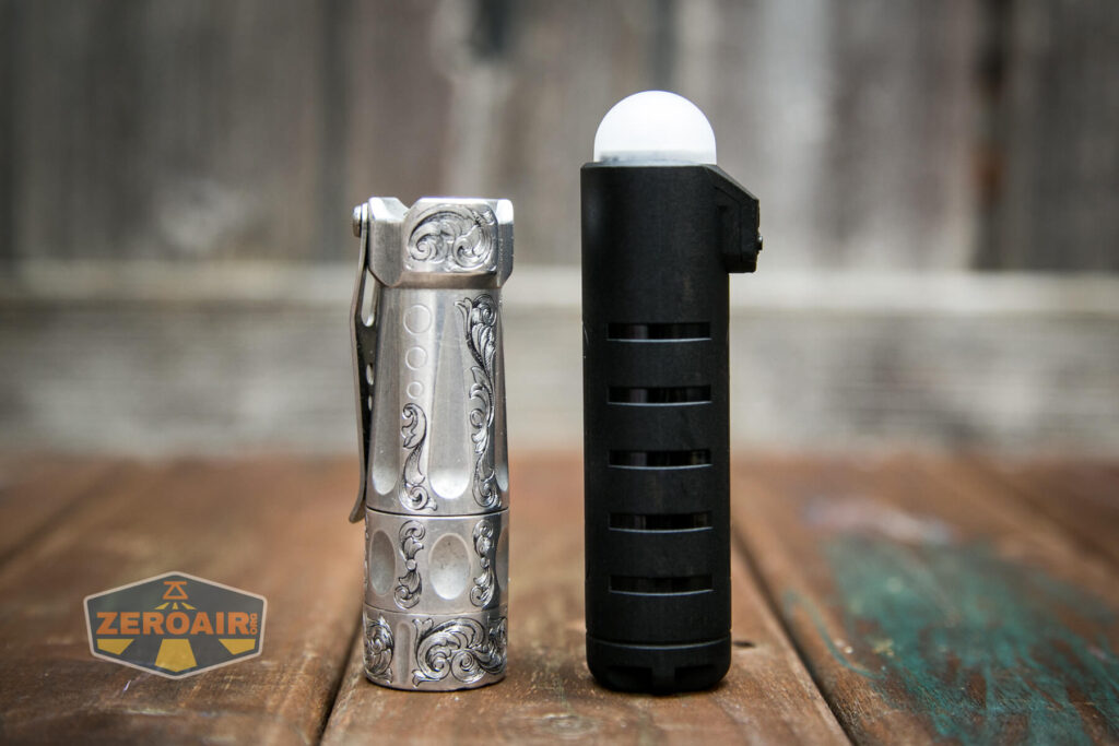 Two handheld flashlights stand upright on a wooden surface. The left flashlight is silver with engraved patterns; the right is black, cylindrical, and topped with a white dome. A ZeroAir logo is visible in the lower left corner.