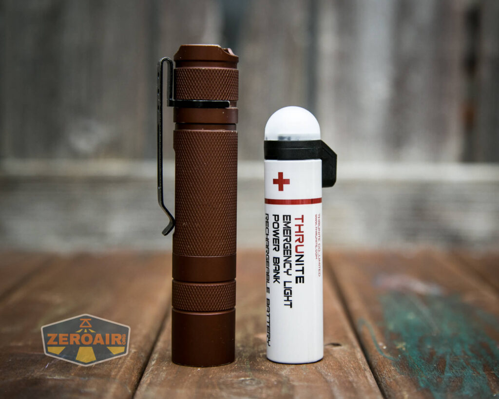 A brown flashlight with a clip stands upright next to a white emergency light and power bank on a wooden surface. The background is blurred, and a ZeroAir logo is visible in the lower left corner.