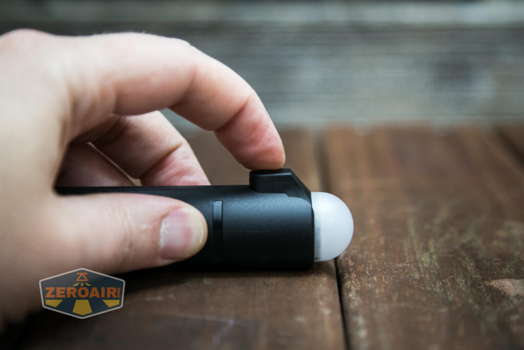 A hand pressing the button on a small black flashlight with a rounded white top, resting on a wooden surface. The ZeroAir logo is visible in the lower left corner.