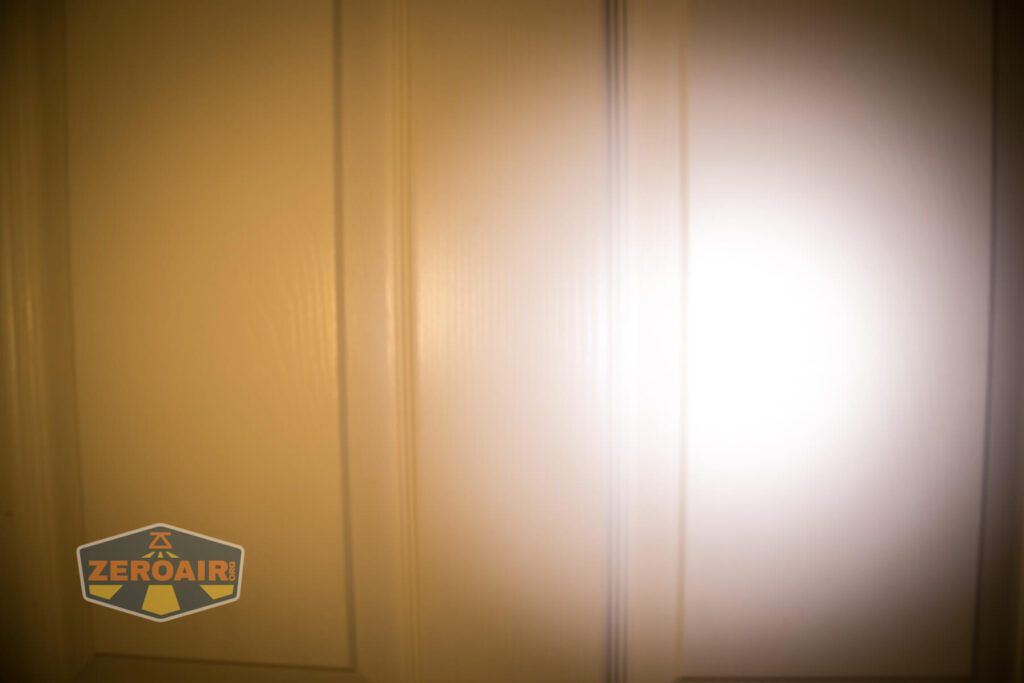 A bright flashlight beam shines on a beige paneled door, creating a circular area of illumination. The lower left corner features a ZEROAIR logo.