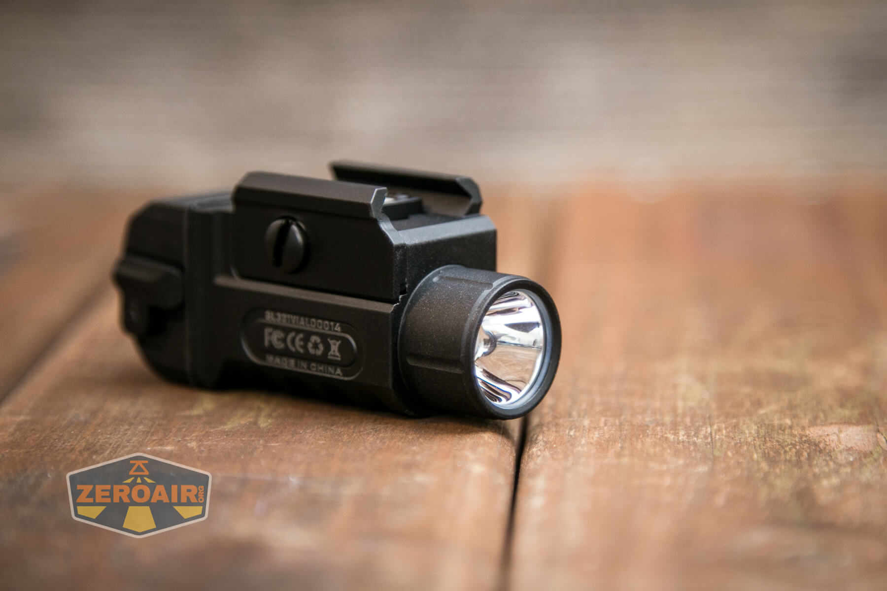 A compact black tactical flashlight with a mounting bracket is placed on a wooden surface. The flashlight is turned off and its lens faces the camera. The ZeroAir logo is visible in the lower left corner.