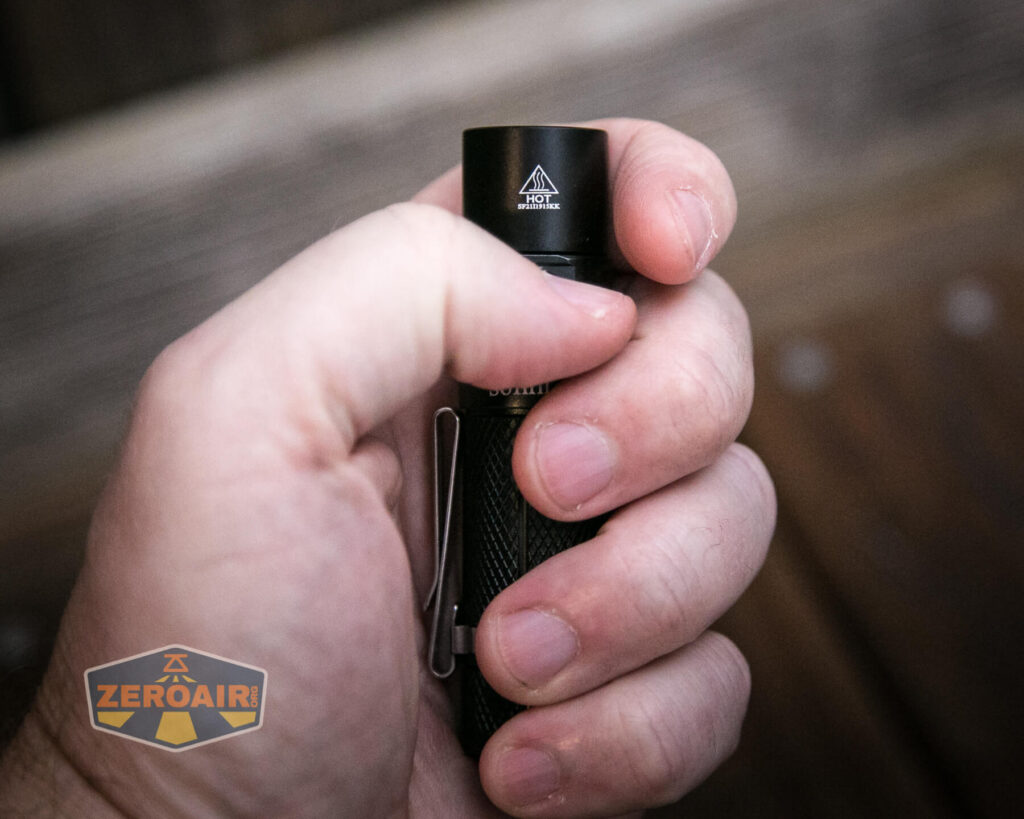 A close-up of a hand holding a small black flashlight with a HOT warning symbol near the top. The flashlight has a textured grip and a clip. The background is blurred and a ZeroAir watermark is visible in the lower left corner.