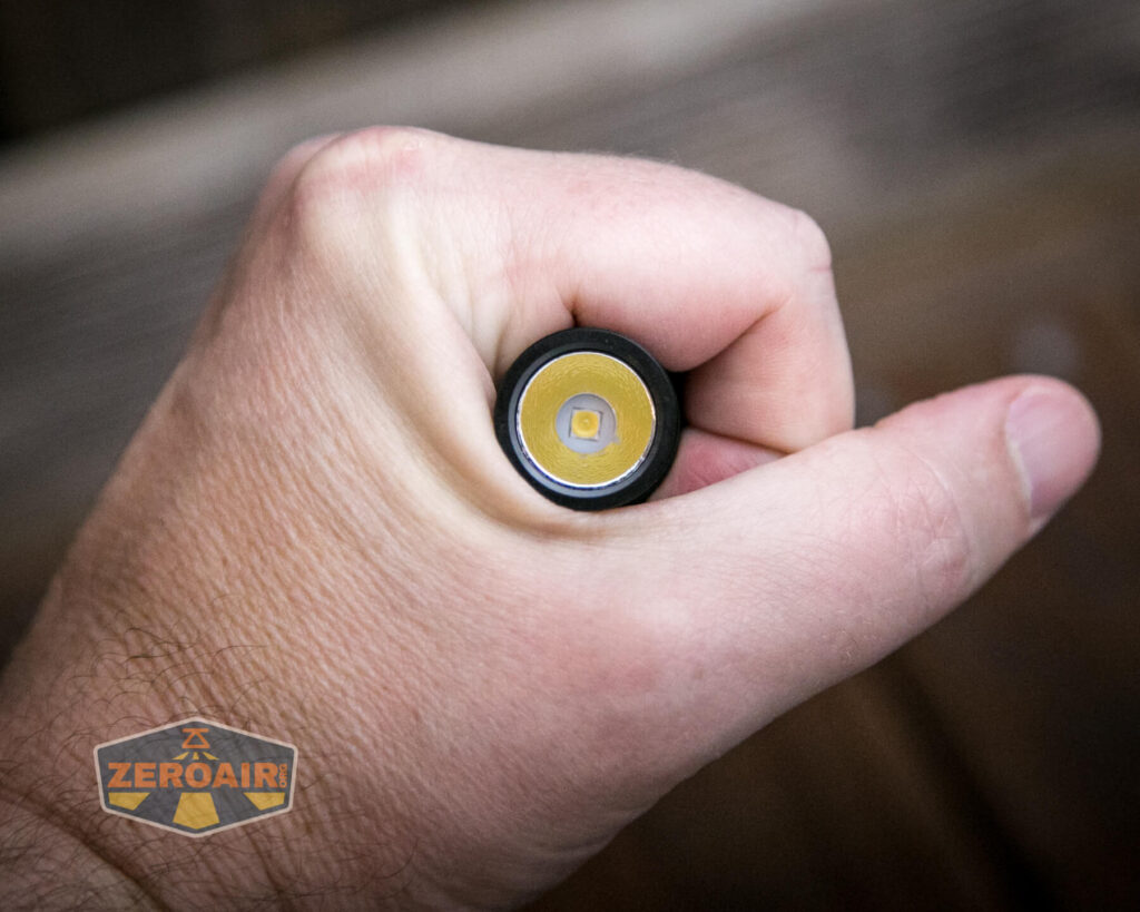 A hand holding a cylindrical battery with the positive terminal facing the camera. The positive terminal has a yellow ring around a silver center. The image includes a ZeroAir logo in the bottom left corner.