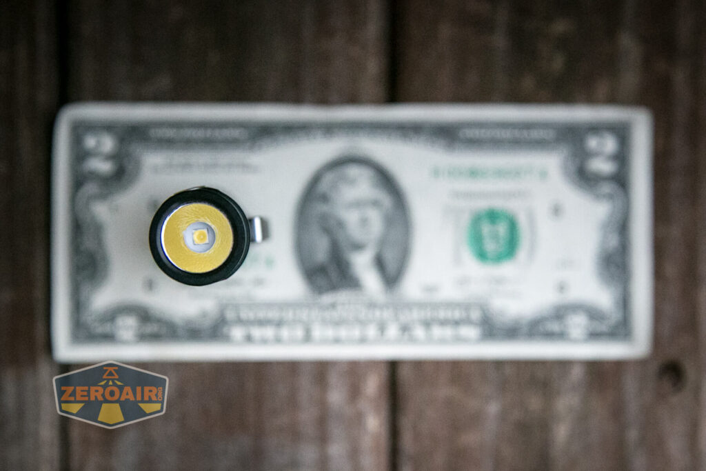 A flashlight head is shown from above, positioned in front of a blurred two-dollar bill on a wooden surface. A logo with the text ZEROAIR is visible in the bottom left corner.