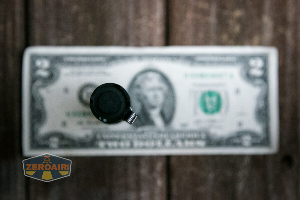 A small cylindrical black object is positioned vertically in the foreground, with the top in focus. Behind it is a blurred U.S. two-dollar bill laying flat on a wooden surface. A ZeroAir logo appears in the lower left corner.