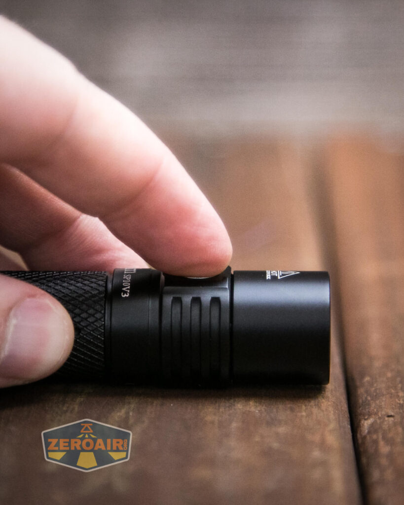 A close-up of a person’s hand pressing the button on a small black flashlight placed on a wooden surface, with a ZeroAir logo in the lower left corner.