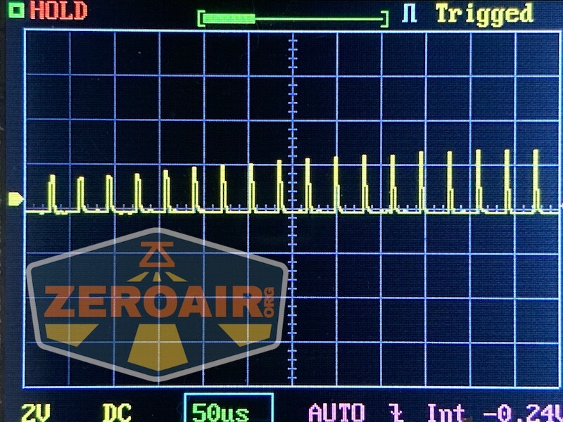 A digital oscilloscope screen displays a repeating square waveform with vertical yellow spikes. The status indicators read HOLD and Triggered, with grid lines and settings shown at the bottom and top left.