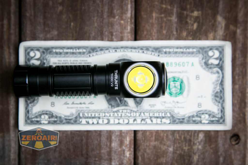 A black Thrunite flashlight lies horizontally on top of a U.S. two-dollar bill. The flashlight’s lens and branding are visible, and a ZeroAir logo appears in the lower left corner of the image.