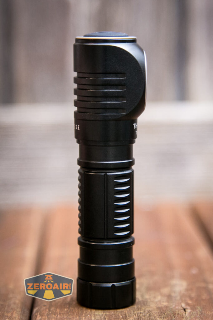 A black flashlight stands upright on a wooden surface with a blurred background. The flashlight has grooves for grip and a button on top. A ZeroAir! logo appears in the bottom left corner.