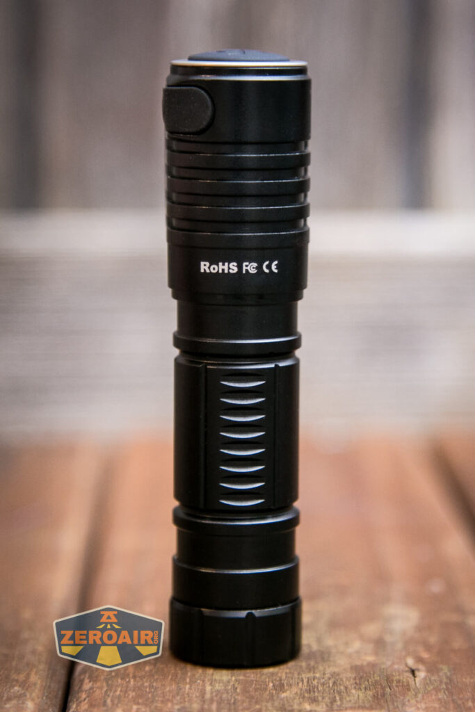 A black cylindrical flashlight stands upright on a wooden surface with a blurred wooden background. The flashlight displays RoHS FC CE markings. A ZEROAIR logo is visible in the lower left corner.