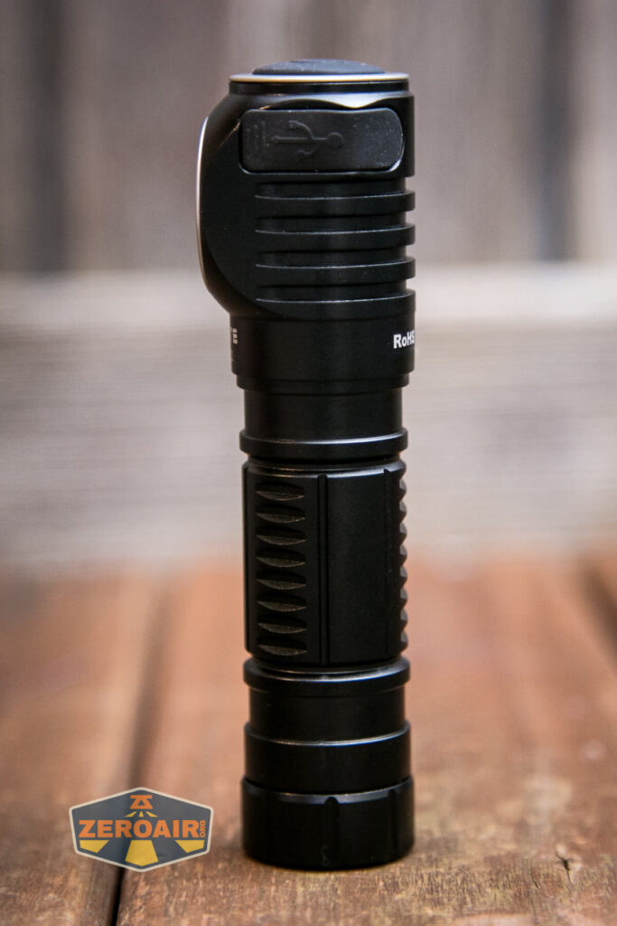 A black flashlight stands upright on a wooden surface with a blurred background. The side of the flashlight shows textured grip patterns and an icon near the top. A ZeroAir logo appears in the lower left corner.