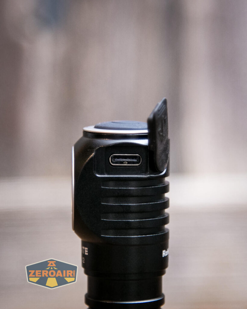 A close-up of a black electronic device with a ribbed grip, showing a USB-C charging port covered by a partially open rubber flap. The background is blurred, and a ZEROAIR! logo appears in the bottom left corner.