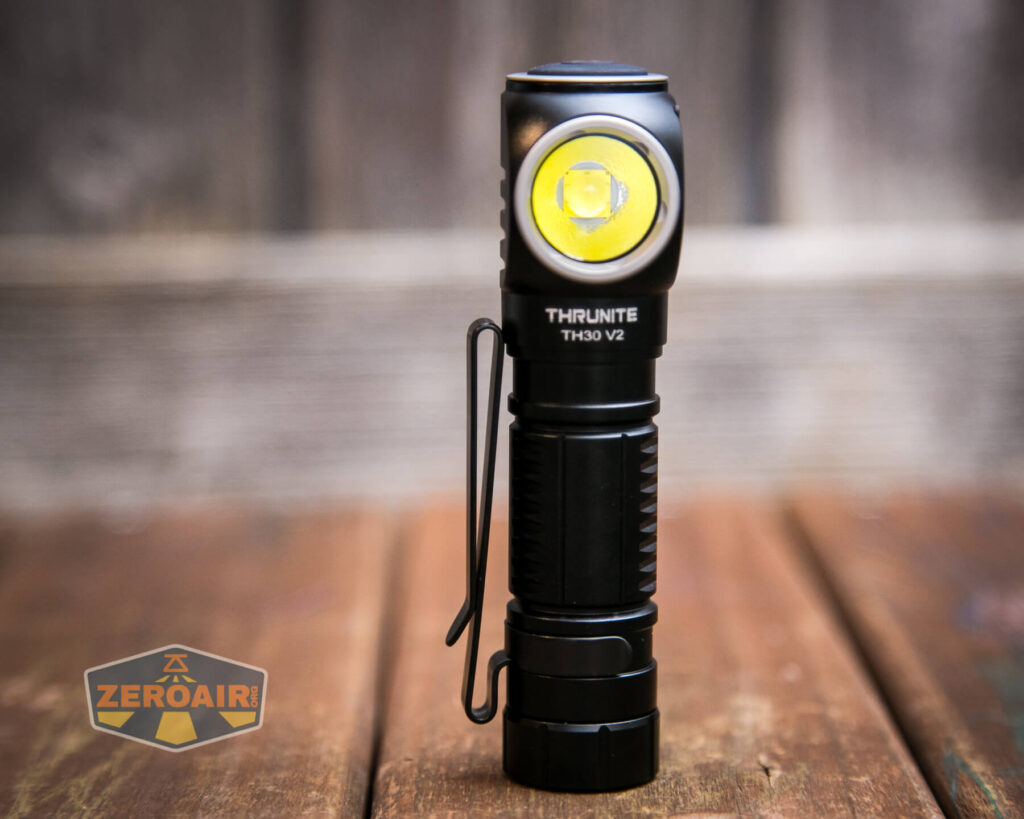 A black ThruNite TH30 V2 flashlight with a side-mounted LED is standing upright on a wooden surface with a blurred wooden background. A ZeroAir logo is visible in the bottom left corner.
