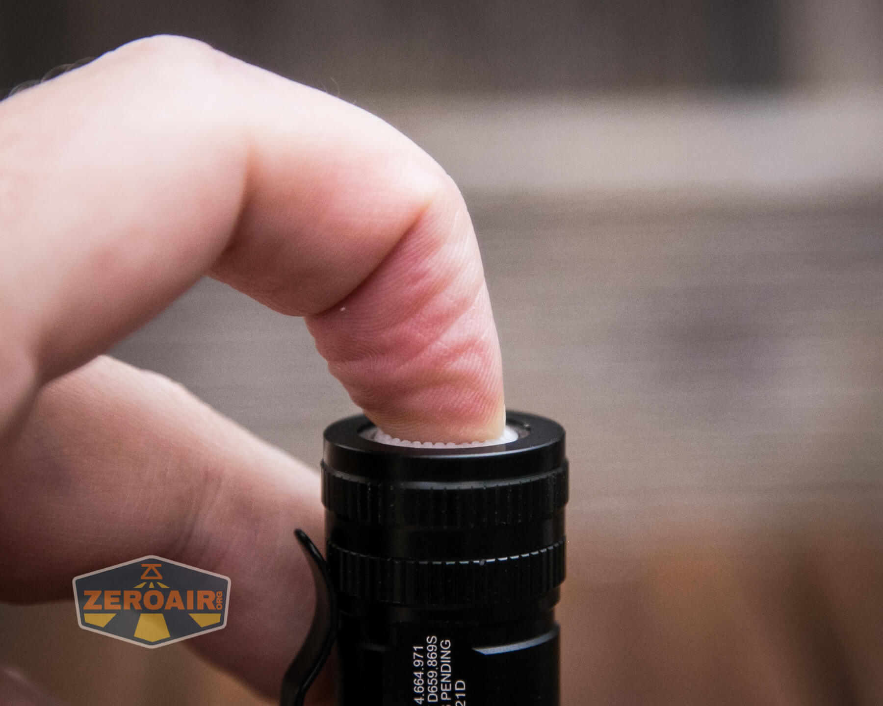 A close-up of a persons finger pressing the button on the end of a black flashlight. The flashlight has a textured grip and a clip attached to its side. The background is blurred.